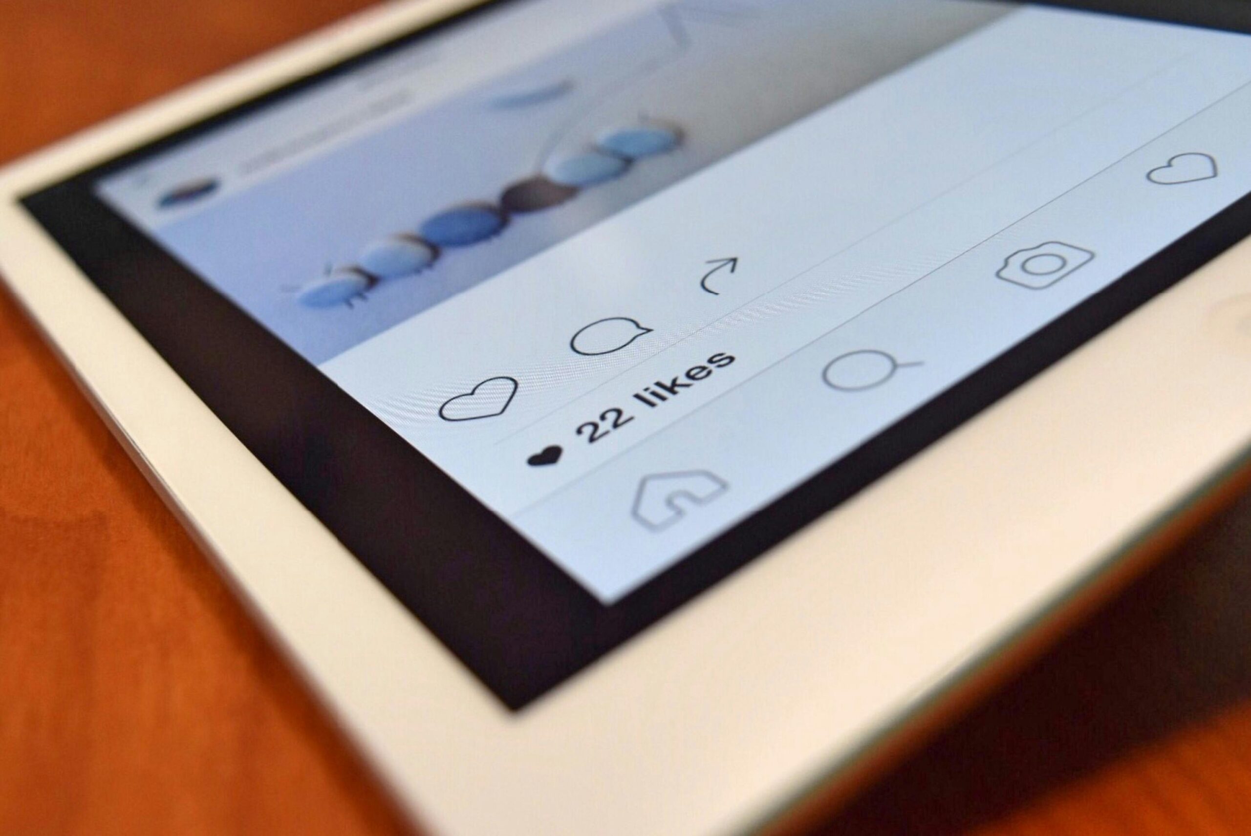 Grande plano de um ecrã de tablet que apresenta uma publicação do Instagram com 22 gostos. A interface mostra ícones para gostar, comentar e partilhar numa superfície de madeira.